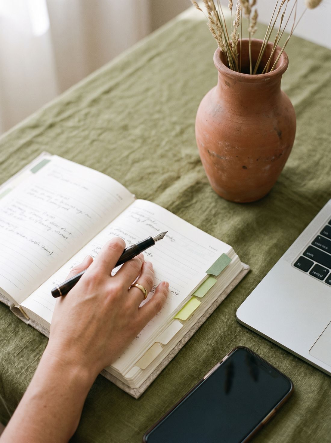 Mão com caneta sobre uma agenda semanal organizada, ao lado de celular e notebook